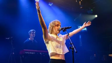 Eine junge Frau hat die Arme ausgebreitet und singt in ein Mikrofon.