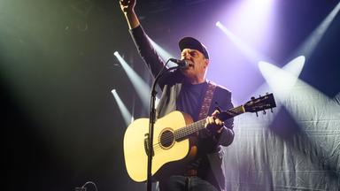 Ein Mann mit Gitarre und singt in ein Mikrofon. Seine Faust ist in die Luft gereckt.