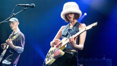 Eine Frau mit Hut am Mikro und E-Gitarre in der Hand auf einer Bühne 