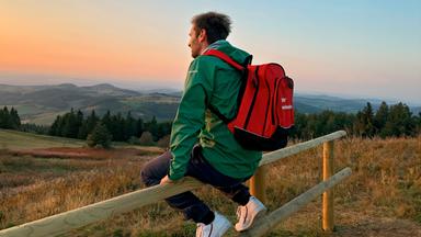 Ein Mann sitzt auf einem Zaun und beobachtet Sonnenaufgang.