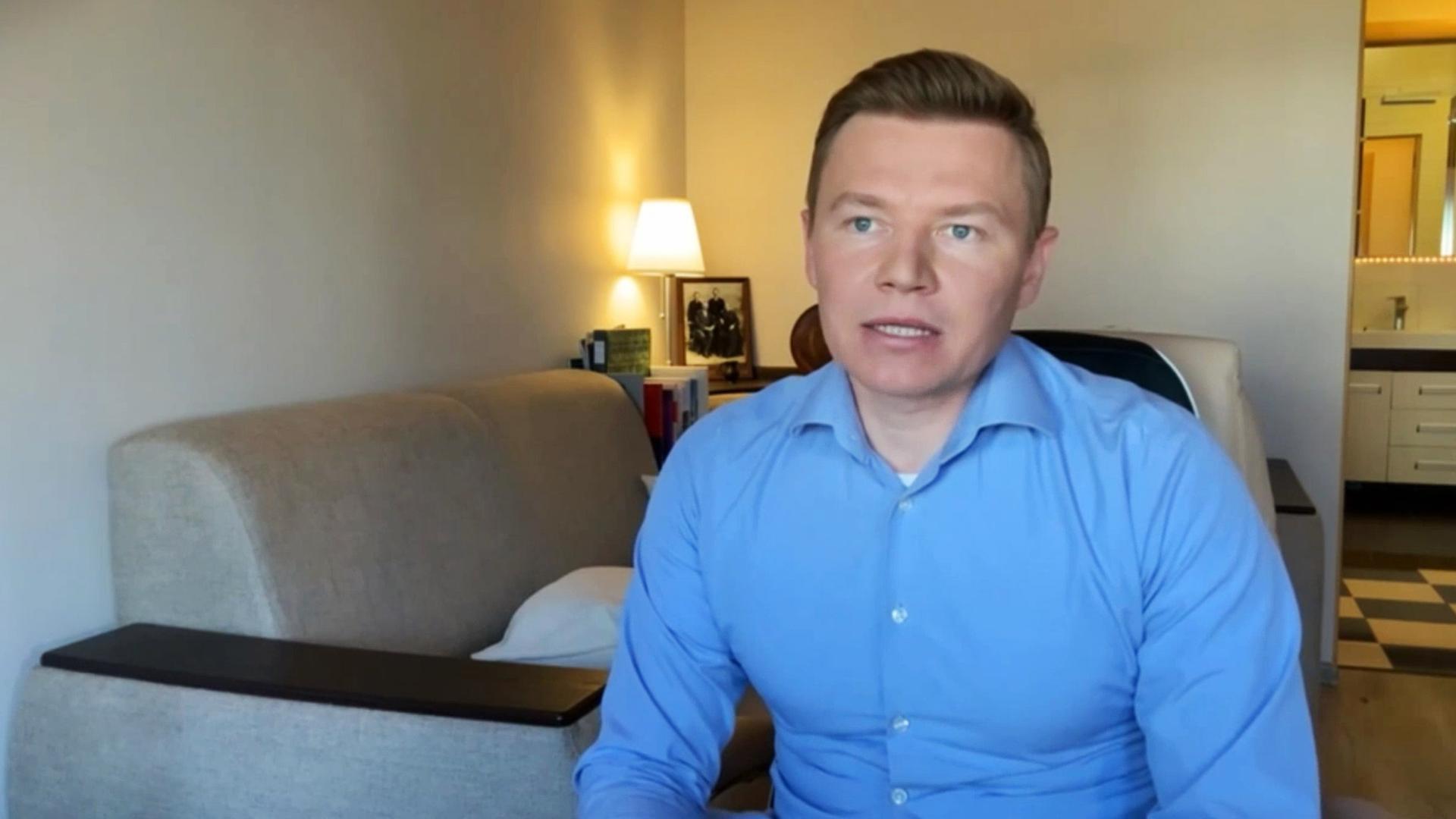 Zu sehen ist ein Mann im blauen Hemd, der in einem Raum mit Sofa und offener Badtür sitzt und leicht den Mund geöffnet hat.