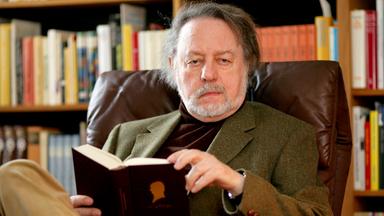 Ein Mann sitzt vor einem Bücherregal und hält ein schwarzes Buch in seiner Hand.
