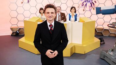 70er Jahre: Hans Rosenthal steht im schwarzen Anzug vor einer Wabenwand im Studio. Hinter ihm sitzt seine Ratejury, Mady Riehl (Theresia Wald), "Ekki" Fritsch (Timo Dierkes) und Brigitte Xander (Katharina Völkl), an Pulten.