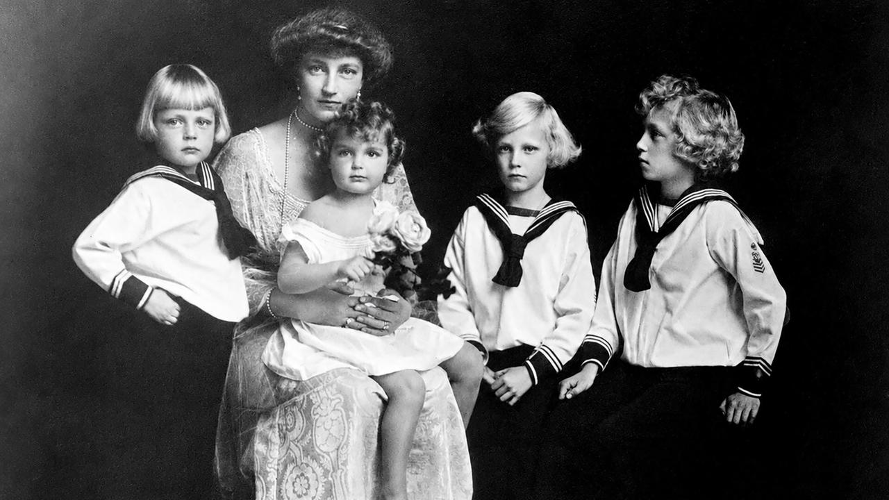 Das Bild zeigt eine Frau mit langen, lockigen Haaren, die ein Kind im weißen Kleid auf dem Schoß hält. Um sie stehen drei weitere Kinder in maritimer Kleidung. Der Hintergrund ist dunkel.