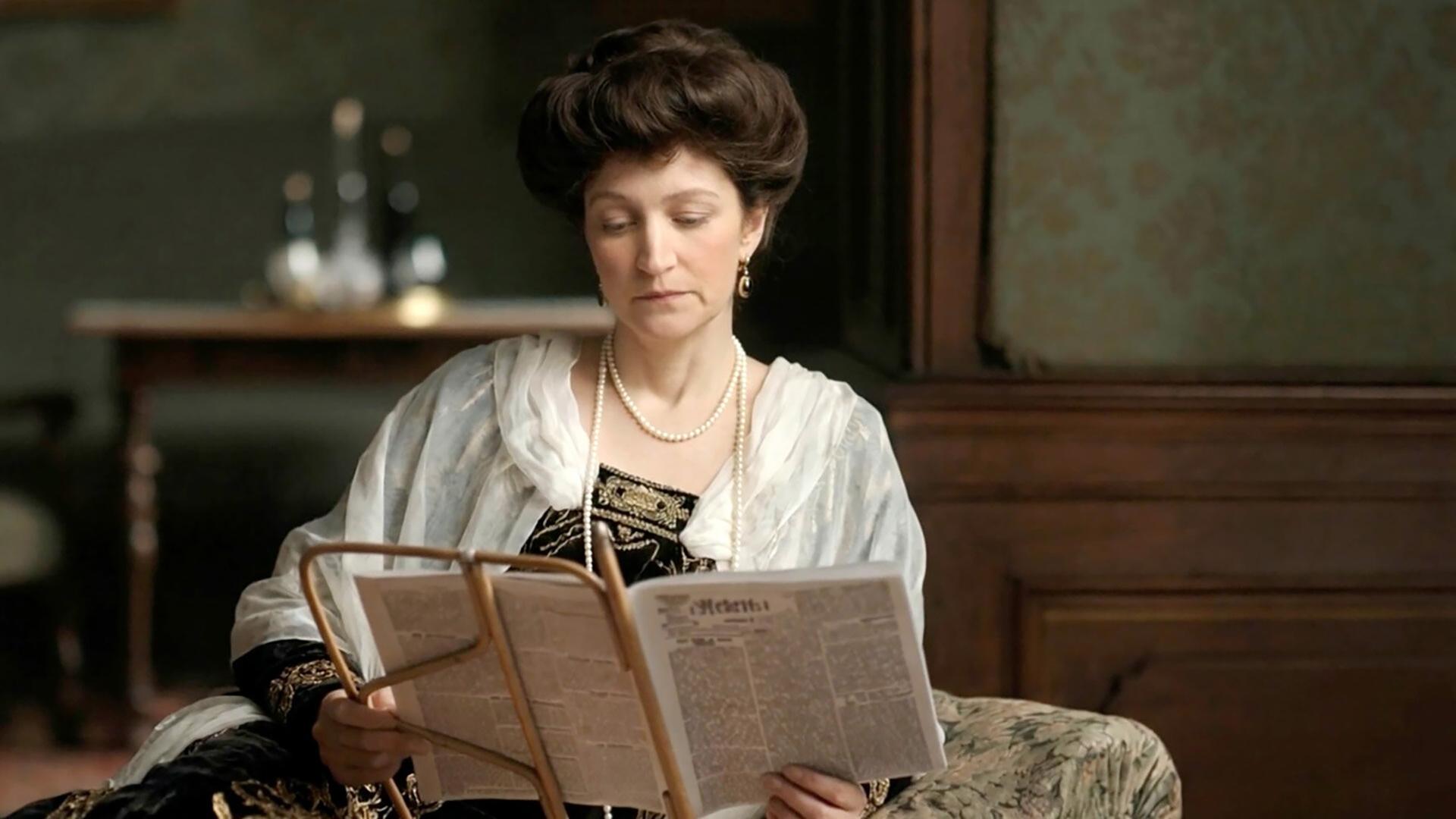 Die Abbildung zeigt eine Frau in historischer Kleidung, die in einem Raum sitzt und eine Zeitung liest. Der Raum ist mit Holzvertäfelungen und einer blauen Tapete dekoriert.