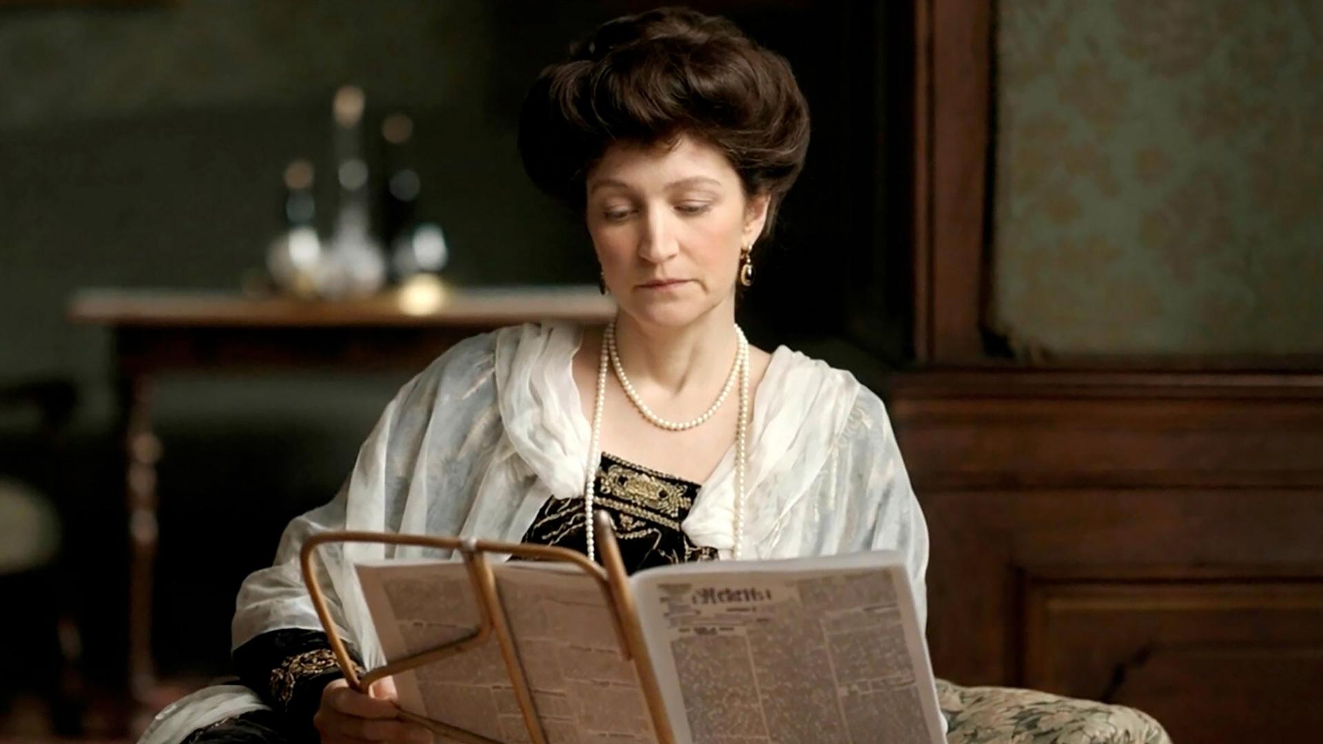Die Abbildung zeigt eine Frau in historischer Kleidung, die in einem Raum sitzt und eine Zeitung liest. Der Raum ist mit Holzvertäfelungen und einer blauen Tapete dekoriert.