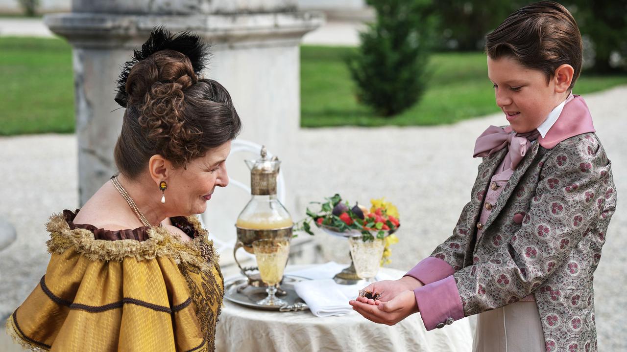 Das Bild zeigt eine Szene aus dem Doku-Drama „Die Rothschild-Saga“. Im Vordergrund sind zwei Personen zu sehen: eine Frau und ein Junge.   Die Frau trägt ein prunkvolles, gelbes Kleid mit einer auffälligen Kragenpartie und hat lockiges, braunes Haar, das zu einem aufwändigen Haarknoten frisiert ist. Sie lächelt und schaut den Jungen an.   Der Junge trägt einen eleganten, hellen Anzug mit einem gemusterten Jackett und einer großen, rosa Schleife um den Kragen. In seiner Hand hält er ein kleines Objekt und präsentiert es der Frau.  Im Hintergrund ist ein Tisch mit einem silbernen Gefäß und Obst in einer Schüssel zu sehen. Die Szenerie scheint im Freien zu sein, umgeben von grünem Gras und Bäumen.   Die gesamte Komposition vermittelt eine gediegene, historische Atmosphäre, die zum Thema des Films passt.