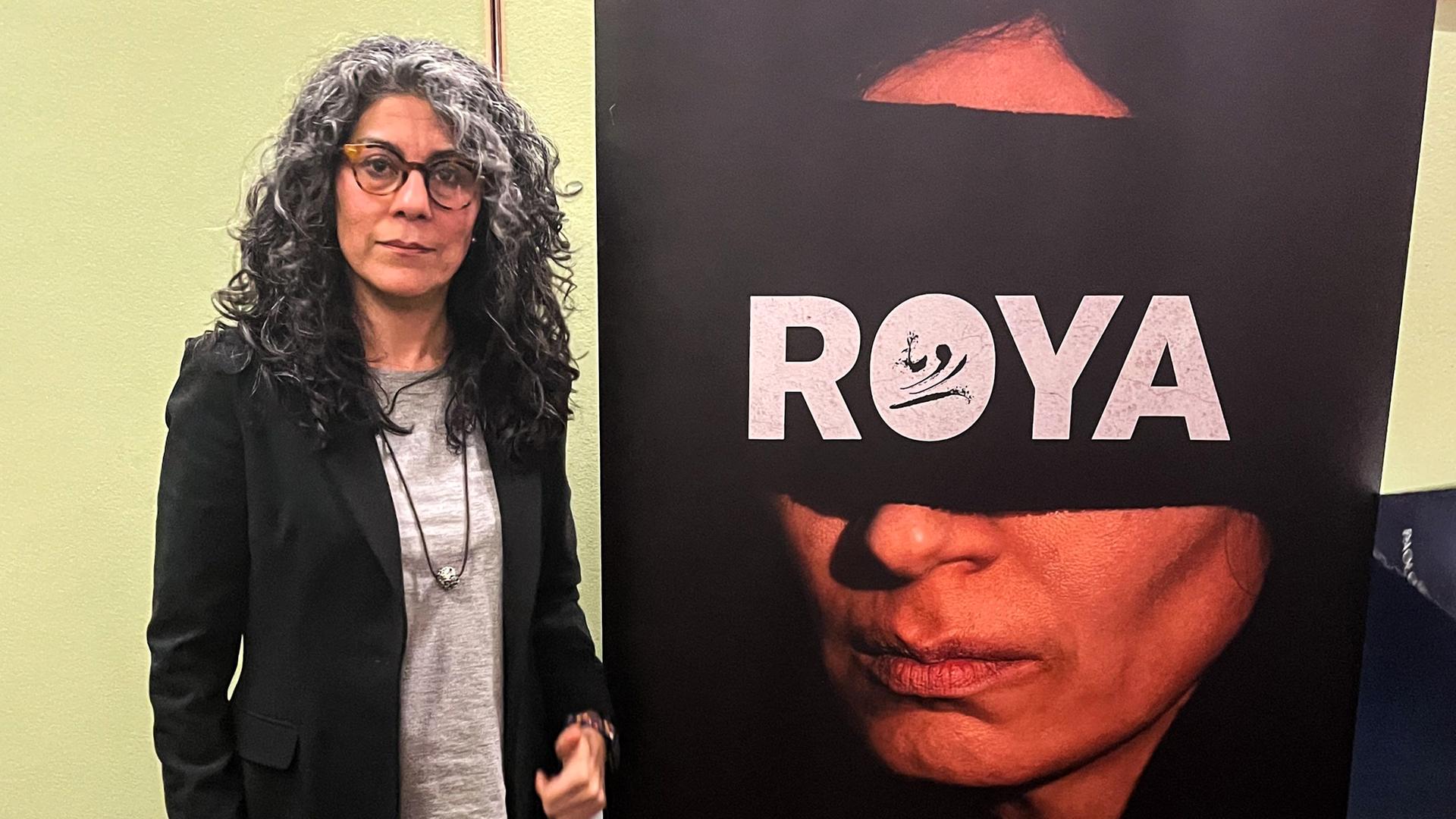 Eine Frau mit langem lockigen und graumeliertem Haar steht neben einem Filmplakat, das ein Frauengesicht mit einer breiten schwarzen Augenbinde zeigt, auf der Roya in Weiß geschrieben steht.
