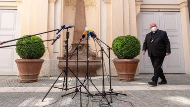 Ein älterer Mann im Anzug mit einer Maske, der in Richtung von mehreren Mikrofonen läuft.