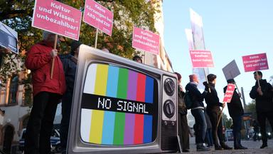 Menschen halten Schilder hoch, auf einer Fernsehattrappe ist "No Signal" zu lesen.