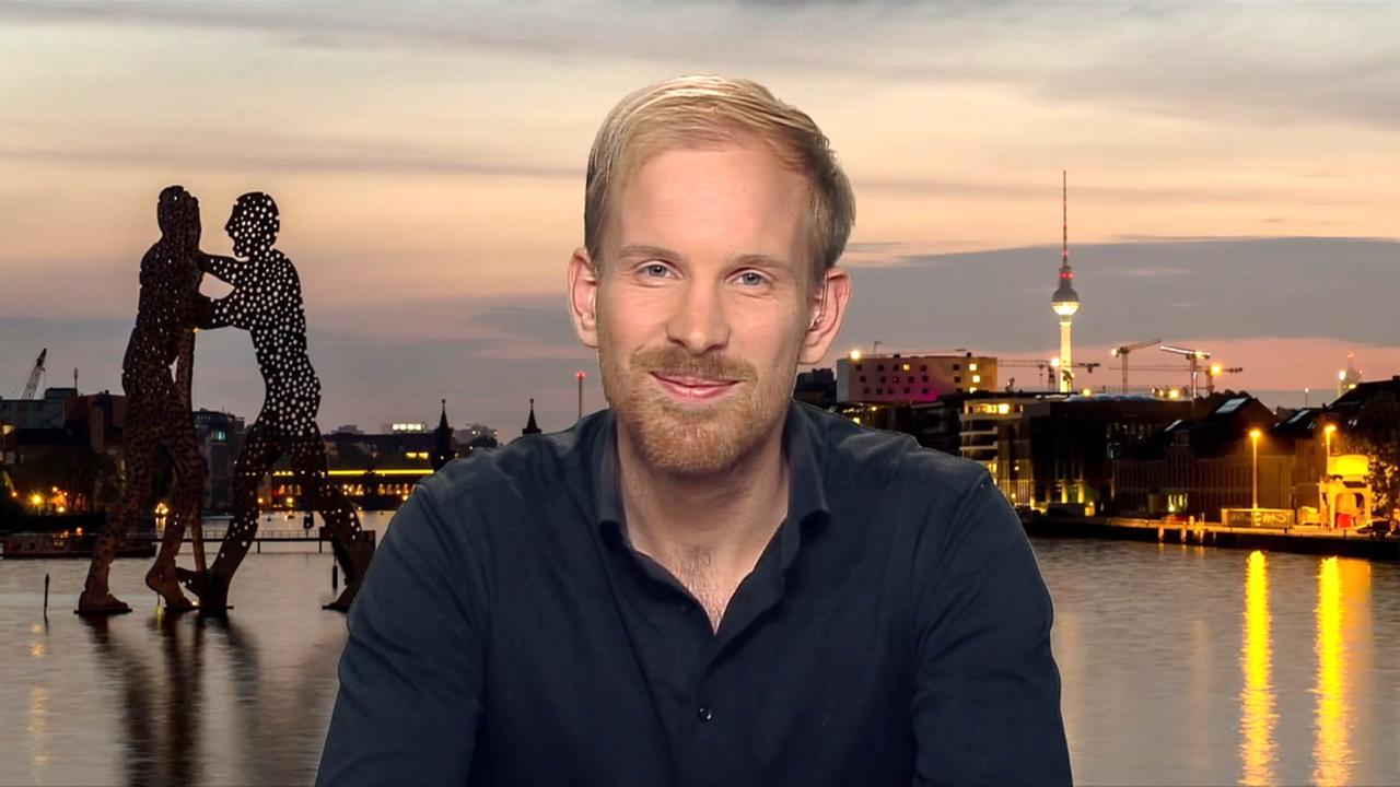 Mann mit blonden Haaren und dunklem Hemd sitzt vor einem Hintergrundbild, das einen See und eine Stadt zeigt.