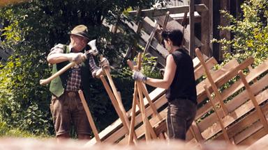 Zwei Menschen bauen mit Äxten einen traditionellen Pinzgauer-Zaun vor einem alten Holzhaus im Grünen.