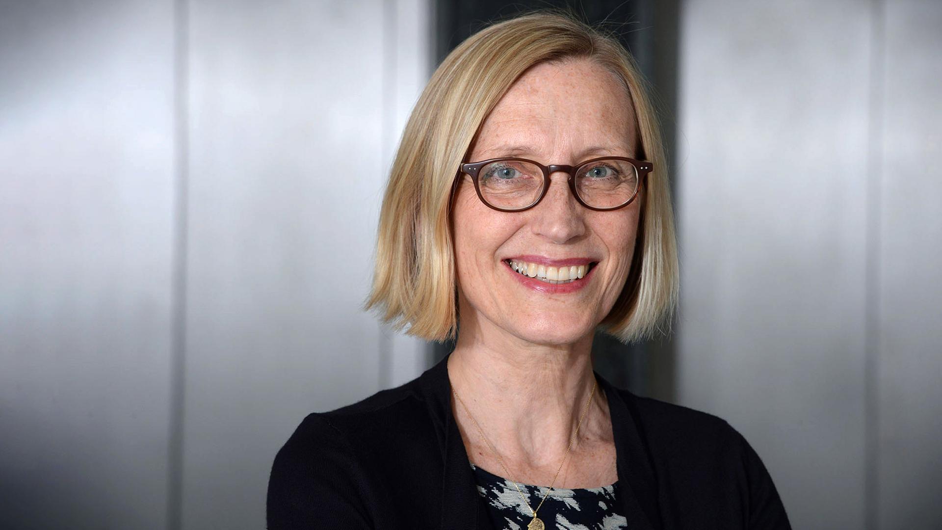 Eine Frau mit halb langem blondem Haar und Brille lächelt.