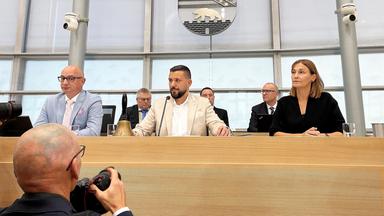 Hinter einem holzverkleideten Podest sitzen zwei Männer und eine Frau, hinter ihnen ist eine weitere Reihe mit Männern in Anzügen zu sehen. Über allen hängt das Wappen von Sachsen-Anhalt.