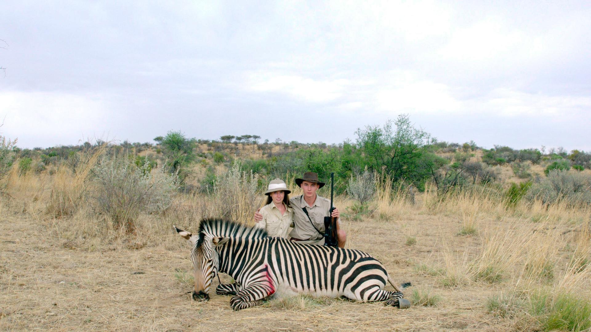 Das Bild zeigt zwei Personen, die in der afrikanischen Savanne vor einem erlegten Zebra posieren. Die Umgebung ist grasbewachsen und es gibt Sträucher. Die Personen tragen Safari-Kleidung und halten ein Gewehr.