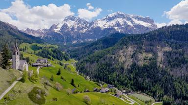 Das Bild zeigt eine atemberaubende Landschaft in Südtirol. Im Vordergrund steht eine kleine, weiße Kirche mit einem spitzen Turm, die auf einem sanften Hügel thront. Die Uhr am Turm ist deutlich sichtbar. Rund um die Kirche erstreckt sich eine weitläufige, grüne Wiese mit vereinzelt stehenden Bäumen.  Im Hintergrund erheben sich beeindruckende, schneebedeckte Berge, deren Gipfel schroff und zerklüftet sind. Diese Berge sind von einem sattgrünen Wald umgeben, der sich bis zu den Bergflanken zieht. Der Himmel ist überwiegend blau mit einigen weißen Wolken, die die sonnige Atmosphäre unterstreichen.  Auf den sanften Hügeln sind einige kleine Siedlungen zu erkennen, die aus traditionellen, mehrgeschossigen Gebäuden bestehen. Die Straßen sind geschwungen und ziehen sich durch die Landschaft. Insgesamt vermittelt das Bild eine starke Verbindung zwischen Natur und menschlicher Siedlung in dieser malerischen Region.