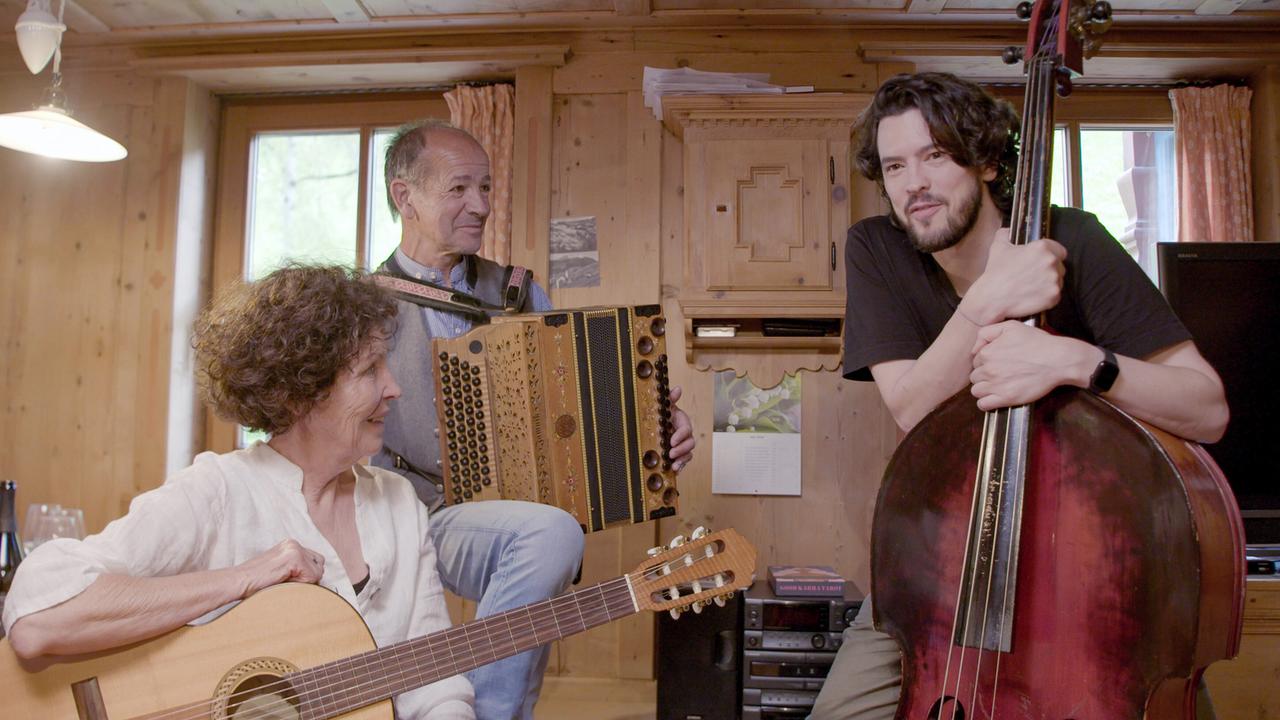 Das Bild zeigt eine gemütliche, holzvertäfelte Innenraumszene. Im Zentrum sind drei Personen versammelt, die musizieren.   - Eine Frau mit lockigen, offenen Haaren sitzt auf der linken Seite und hält eine akustische Gitarre. Sie trägt eine weiße Bluse und lächelt. - In der Mitte steht ein Mann, der ein Akkordeon spielt. Er hat kurzes Haar und trägt ein hellblaues Hemd sowie eine graue Weste.  - Auf der rechten Seite steht ein junger Mann mit langen, dunklen Haaren. Er hat ein schwarzes T-Shirt an und umarmt ein Cello.  Der Raum wirkt hell und einladend, mit einem Fenster im Hintergrund, das Tageslicht hineinlässt. An den Wänden sind einige Bilder und ein Wandregal zu sehen, und es gibt einen kleinen Tisch mit einer Flasche im Vordergrund.  Die Szene vermittelt eine Atmosphäre von Gemeinschaft und musikalischer Tradition.