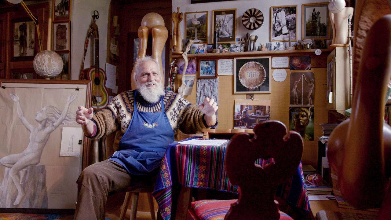 Das Bild zeigt einen älteren Mann mit einem gepflegten, grauen Bart, der in einem kreativen Atelier sitzt. Er trägt einen blauen Latzschürze und ein gestricktes Oberteil mit einem bunten Muster. Der Mann hat eine einladende Körperhaltung und wirkt im Gespräch oder in einer Erzählung vertieft.  Hinter ihm ist eine Wand voller Kunstwerke und Fotografien. Einige Bilder zeigen Skulpturen und Zeichnungen, darunter eine Darstellung einer menschlichen Figur. Auf einem Tisch vor ihm liegt bunte, gestreifte Tischdecke und darauf sind verschiedene Unterlagen und Materialien verstreut.   Die Umgebung wirkt warm und einladend, mit viel Holz und einer Vielzahl von künstlerischen Requisiten, darunter auch eine Lampe und einige Musikinstrumente. Das Licht ist sanft und trägt zur gemütlichen Atmosphäre bei.   Die gesamte Szenerie vermittelt den Eindruck von Kreativität und einer tiefen Verbindung zur Kunst.