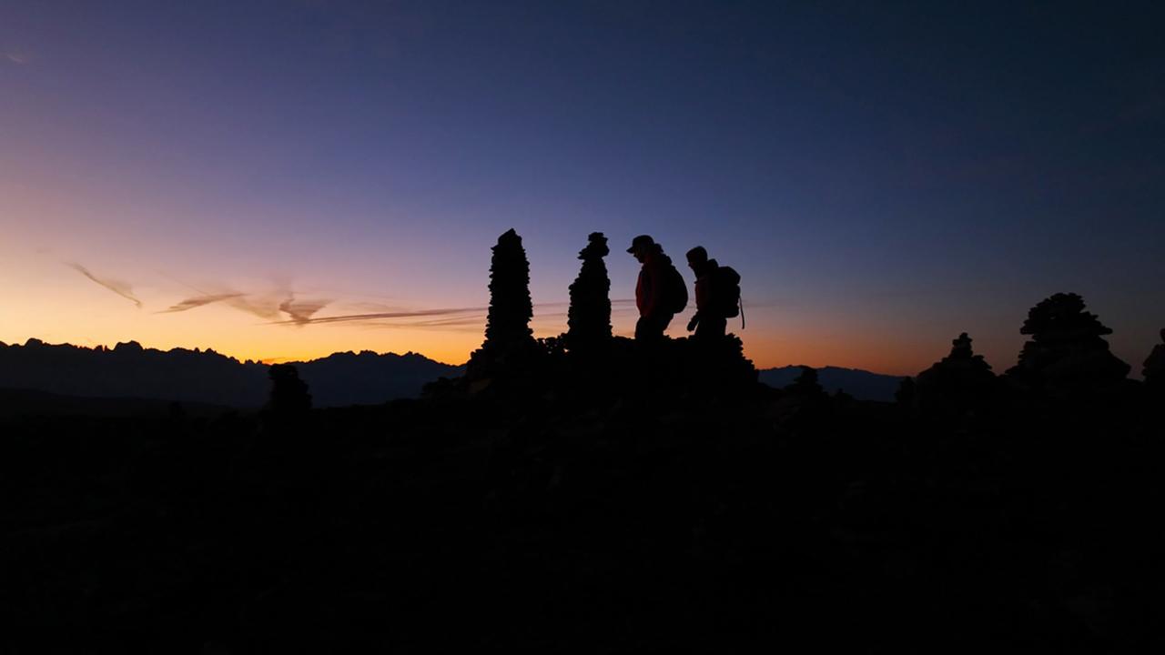 Das Bild zeigt eine silhouettehafte Landschaft bei Sonnenaufgang, die von einem klaren, tiefblauen und orangefarbenen Himmel überlagert wird. Im Vordergrund sind mehrere große Steinfiguren, die übereinander gestapelt sind, als „Stoanerne Mandln“ bekannt. Zwei Wanderer, einer in hellen und einer in dunklen Kleidern, befinden sich rechts im Bild. Die Silhouetten der Wanderer heben sich vom helleren Himmel ab. Im Hintergrund sind die schroffen Konturen der Dolomiten zu sehen, die eine ikonische Berglandschaft darstellen. Der Gesamteindruck vermittelt eine mystische Atmosphäre, die den Zauber und die Legenden der Region Südtirol widerspiegelt.