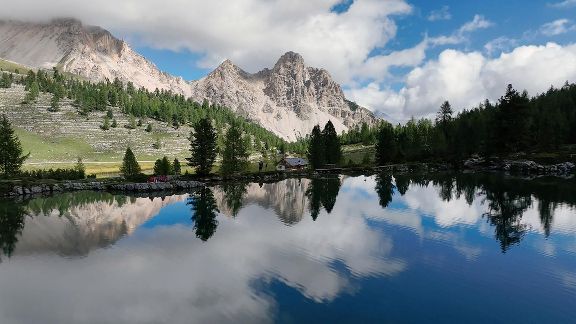 Das Bild zeigt eine malerische alpine Landschaft, die von hohen Berggipfeln umgeben ist. Im Vordergrund ist ein ruhiger Bergsee zu sehen, dessen Wasser die umgebenden Berge und den Himmel reflektiert. Die Wasseroberfläche ist spiegelglatt, wodurch die Wolken und die Gipfel in einer klaren Spiegelung erscheinen.   Rechts am Seeufer steht ein kleines, rustikales Holzhaus, umgeben von einigen Nadelbäumen. In der Nähe des Hauses sind zwei Personen zu erkennen, die auf einem Weg stehen. Die Hügel im Hintergrund sind mit schroffen Felsen und Bäumen bedeckt. Der Himmel ist teilweise bewölkt, mit einigen blauen Stellen, die durch die Wolken sichtbar sind.   Insgesamt vermittelt die Szene ein Gefühl von Ruhe und einer unberührten Naturlandschaft, typisch für die Region Südtirol.
