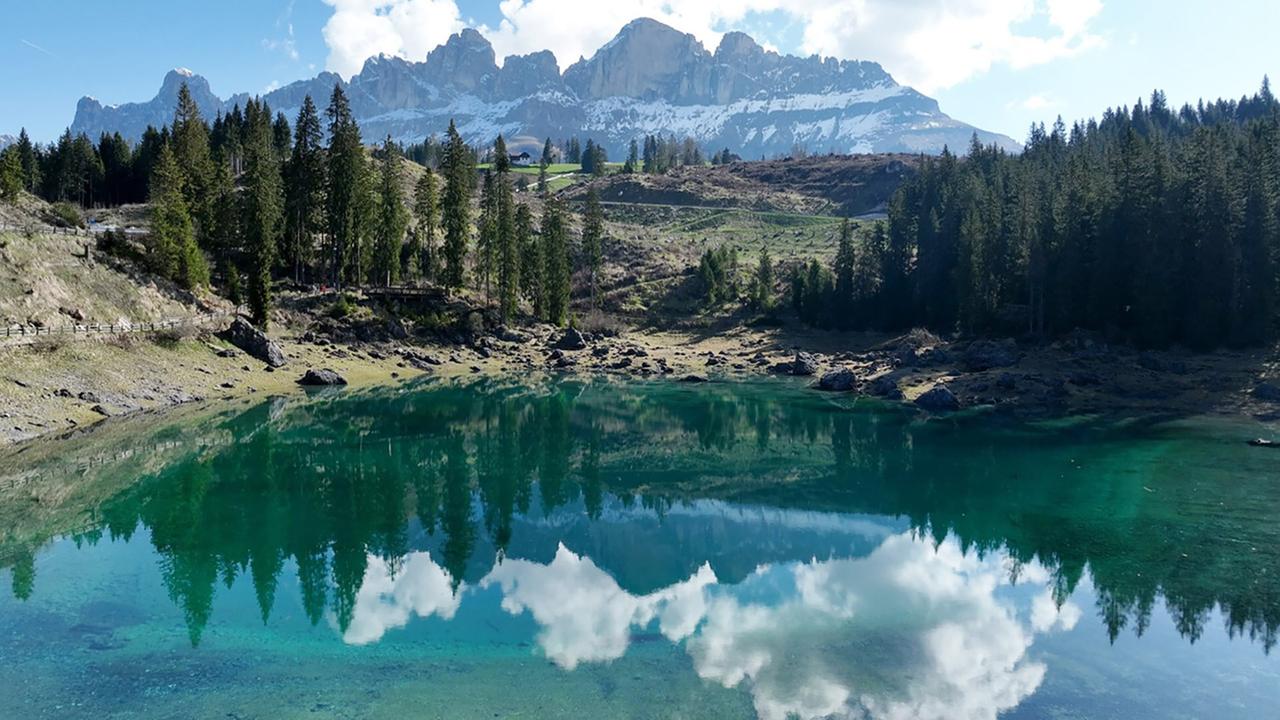 Das Bild zeigt den Karer See in Südtirol, umgeben von majestätischen Bergen der Rosengarten-Gruppe. Der See hat eine klare, türkisfarbene Wasseroberfläche, die die Wolken und umliegenden Bäume spiegelt. Am Ufer des Sees befinden sich einige große Steine und eine schmale, unbefestigte Uferlinie. Darüber hinaus sind hohe Nadelbäume sichtbar, die die Landschaft säumen. Im Hintergrund erheben sich die schroffen, schneebedeckten Gipfel der Dolomiten, die in den Himmel ragen. Die Atmosphäre wirkt friedlich und beeindruckend, mit einem klaren blauen Himmel und vereinzelten Wolken.