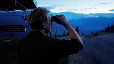 Max Müller beobachtet mit einem Fernglas die Berglandschaft bei Dämmerung. Im Hintergrund sind Berge und ein Sessellift zu sehen.