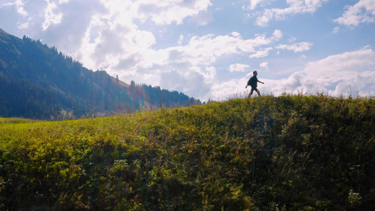 Max Müller läuft über eine Anhöhe in einer sonnigen Berglandschaft. Im Hintergrund sind bewaldete Berghänge und ein blauer Himmel mit Wolken zu sehen.