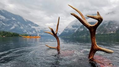 Zwei große Hirschgeweihe ragen aus einem See, während im Hintergrund eine gelbe Plätte mit Menschen darauf fährt. Die Szene spielt vor einer nebelverhangenen Berglandschaft.
