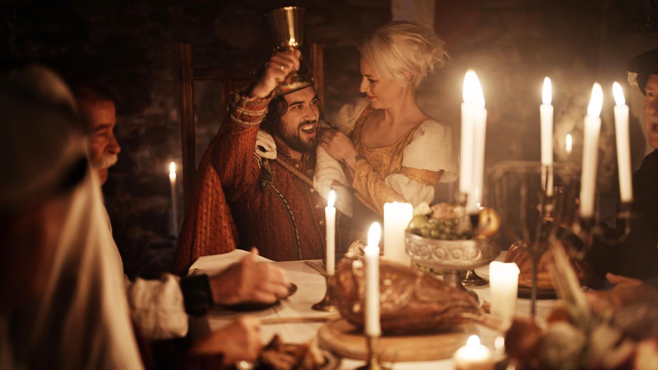 In einem Kerzenschein erleuchteten Raum feiert eine mittelalterlich gekleidete Gesellschaft ein festliches Bankett. Ein Mann mit Krone hebt einen Kelch, während eine Frau ihn anlacht.