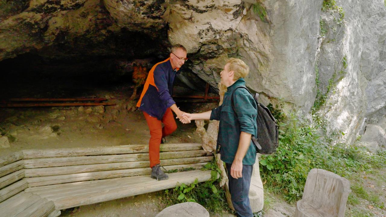 Max Müller und Historiker Michael Kurz stehen vor einer Höhle und schütteln sich die Hände. Müller trägt einen Rucksack und eine grüne Jacke, während Kurz eine Outdoor-Jacke mit orangen Akzenten trägt.