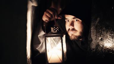 Ein Mann in einer dunklen Kutte hält eine Laterne in einem schmalen, düsteren Gang. Sein Gesicht ist teils im Schatten, während das Kerzenlicht seine Züge beleuchtet.