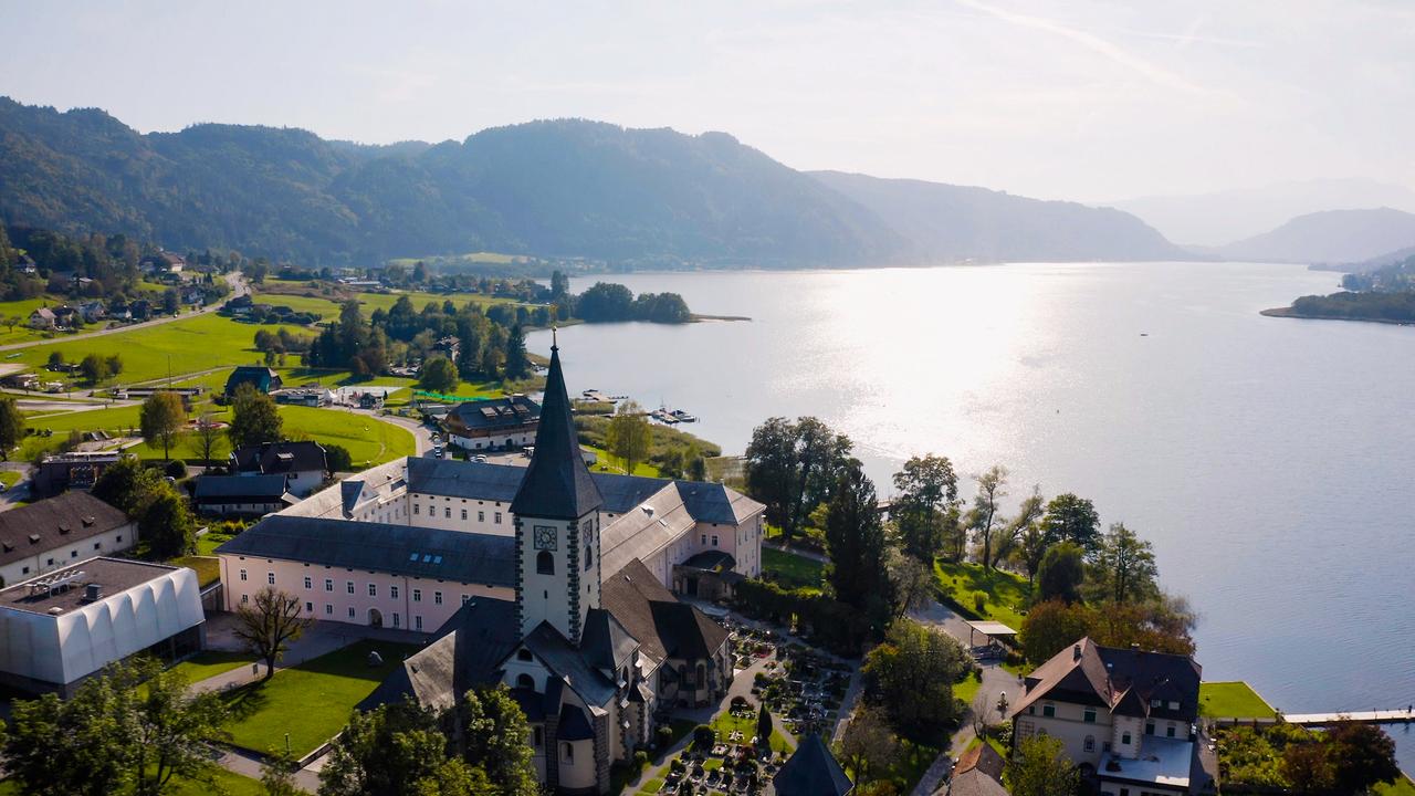 Luftaufnahme des Ossiacher Sees mit einer Klosteranlage im Vordergrund. Der See erstreckt sich in die Ferne, umgeben von grünen Wiesen und bewaldeten Hügeln.