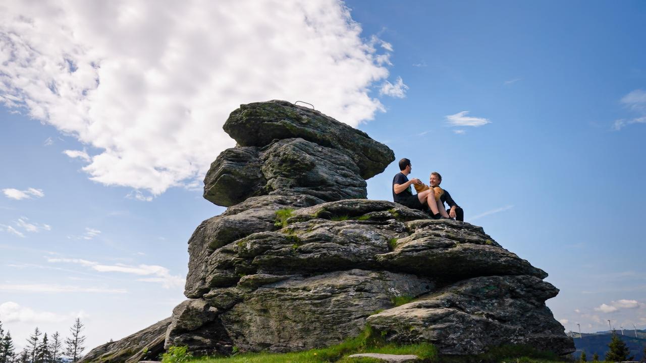 Zwei Männer sitzen auf einem großen, übereinander gestapelten Felsen mit weitem Blick in die Landschaft. Der Himmel ist blau mit weißen Wolken.
