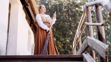Eine Frau in historischer Kleidung steht mit einem Reisigbesen auf einer Holztreppe vor einem alten Haus und blickt nach oben.