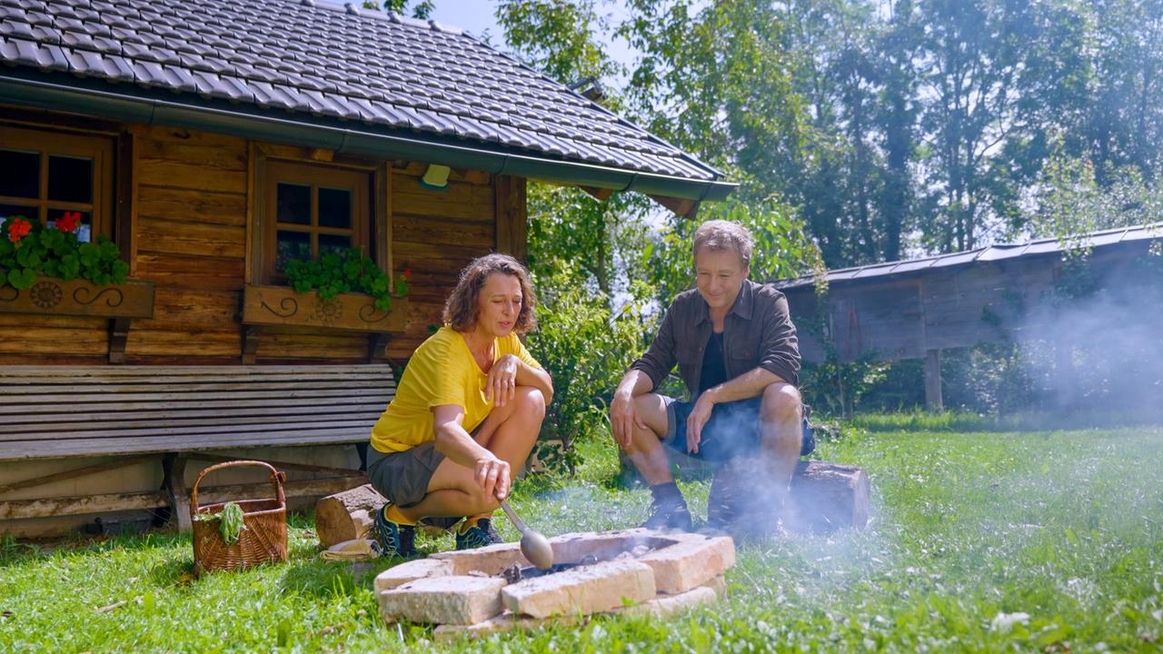 Max Müller und Helga Graef sitzen vor einem kleinen Holzhaus im Grünen und bereiten eine Mahlzeit über offenem Feuer zu.