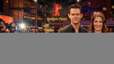 Die "aspekte"-Moderatoren Katty Salié und Jo Schück stehen vor dem Berlinale Palast auf dem roten Teppich. Hinter ihnen ist die Absperrung und das Blitzgewitter der Fotografen sowie das leuchtende Berlinale-Logo zu sehen. Jo Schück hält einen goldenen Bären in der ausgestreckten Hand.