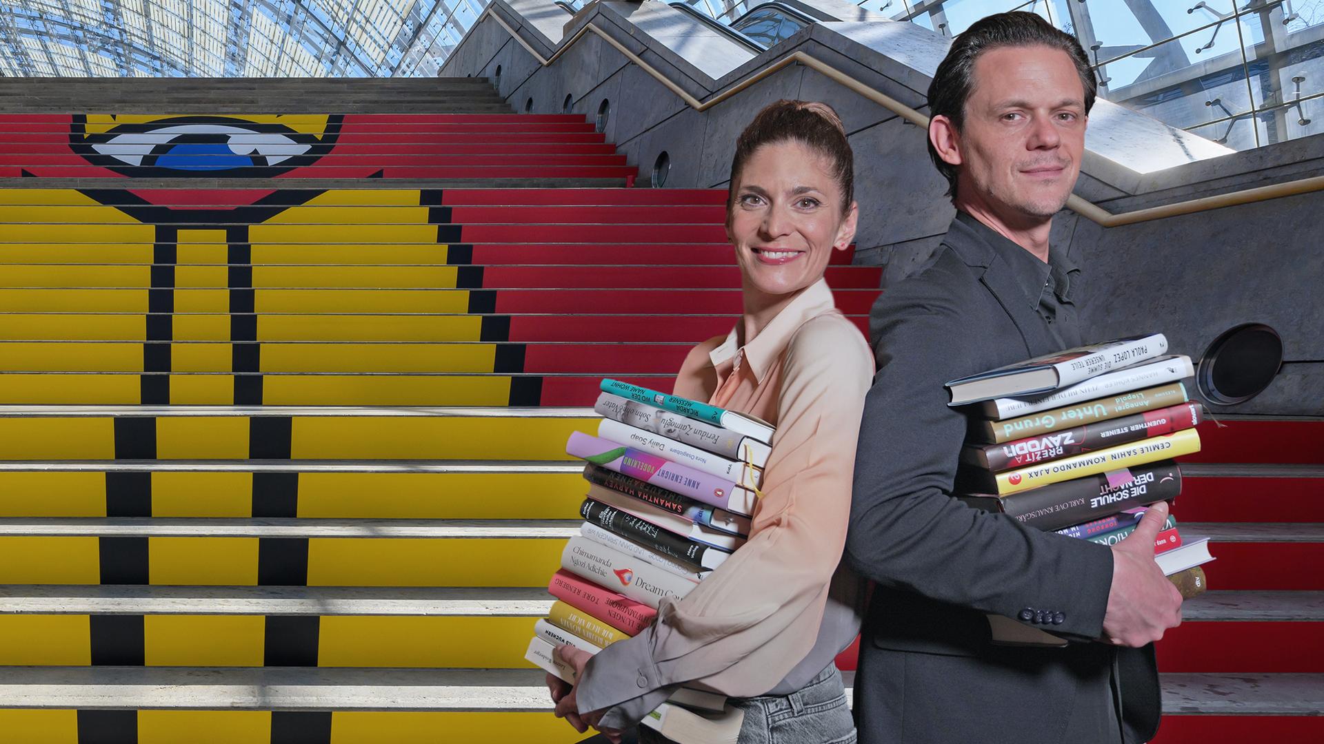 Eine Frau und ein Mann mit Büchern in den Armen stehen in der Ausstellungshalle vor einer Treppe