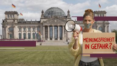 Moderatorin Katty Salié steht vor dem Reichstagsgebäude in Berlin. Sie hält ein Megafon in der rechten Hand und ein Pappschild mit der Aufschrift „Meinungsfreiheit in Gefahr?“ in der linken. Ihr Mund ist mit silbernem Klebeband überklebt.