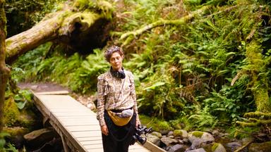 Frau steht auf Brücke in Wald und häklt eine Kamera in der Hand.