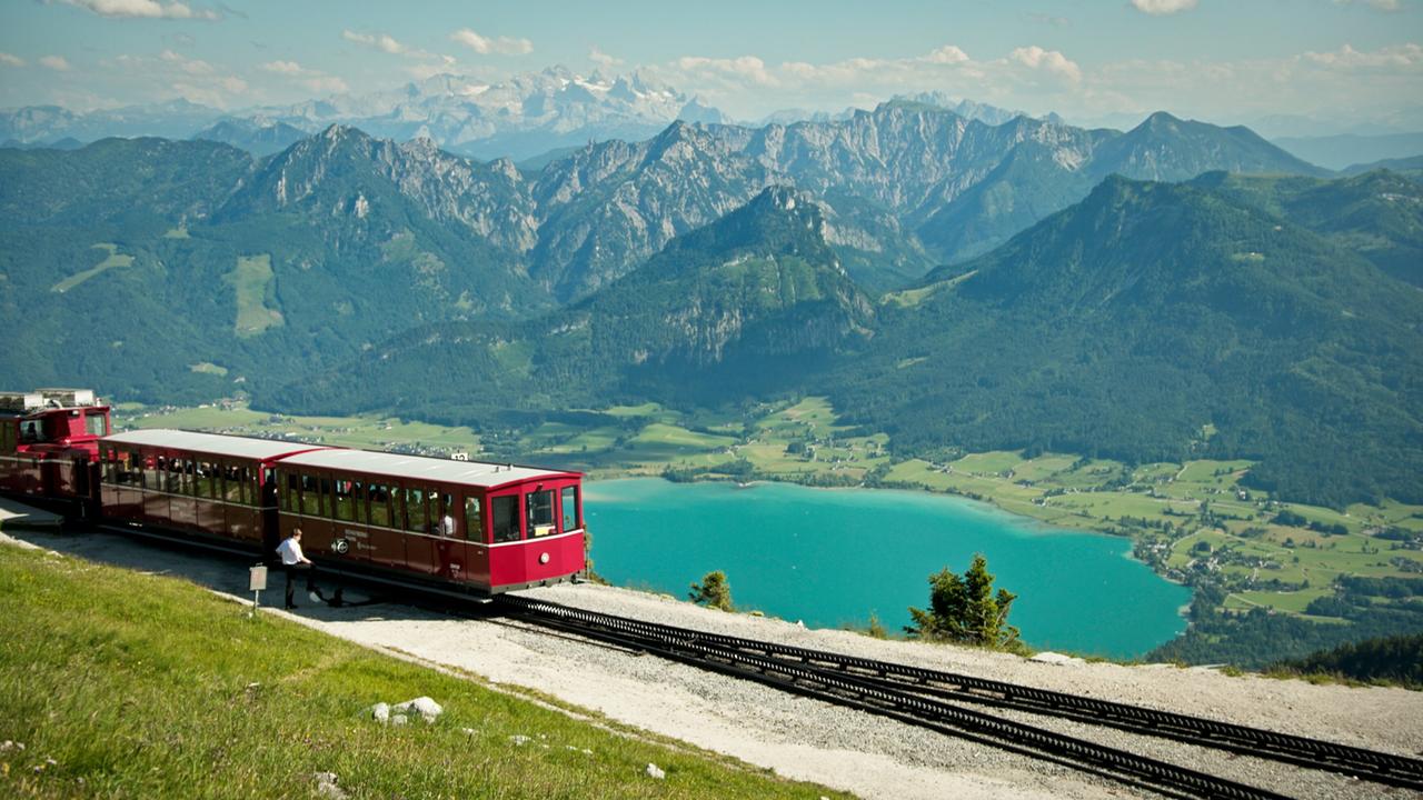 Das Bild zeigt die Schafbergbahn, eine Zahnradbahn, die in den österreichischen Alpen verkehrt. Im Vordergrund ist ein rotes Bahngleis zu sehen, auf dem ein Zug steht. Der Zug hat große Fenster, durch die man die umliegende Landschaft gut erkennen kann.   Hinter dem Zug erstreckt sich eine beeindruckende Berglandschaft mit hohen, bewaldeten Bergen und schroffen Felsformationen. In der Ferne sind schneebedeckte Gipfel sichtbar, was auf eine hohe Lage hinweist.   Zu Füßen der Berge liegt ein großer See mit türkisblauem Wasser, umgeben von grünen Wiesen und kleinen Ortschaften. Der Himmel ist klar mit einigen wenigen weißen Wolken, was auf schönes Wetter hindeutet.   Die gesamte Szenerie vermittelt ein Gefühl von Ruhe und Naturverbundenheit, charakteristisch für das Salzburger Seenland in Österreich.