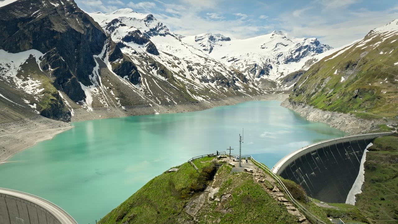 Das Bild zeigt eine beeindruckende Berglandschaft mit einer Staumauer im Vordergrund. Im unteren Teil des Bildes ist ein grüner Hang zu sehen, auf dem sich einige Menschen aufhalten. Rechts im Bild verläuft eine kurvenreiche Straße, die zur Staumauer führt.   Die Staumauer ist groß und hat eine bemerkenswerte, gewölbte Form. Hinter der Staumauer erstreckt sich ein türkisfarbenes Wasserbecken, das von steilen, felsigen Bergen und schneebedeckten Gipfeln umgeben ist. Die Berge sind teilweise mit grünem Gras bedeckt, was einen schönen Kontrast zu den weißen Schneefeldern und dem blauen Himmel bildet.  Der Himmel zeigt einige Wolken, die aber größtenteils lichtdurchlässig sind, sodass das Licht die Szene erhellt. Die Atmosphäre wirkt friedlich und einladend, ideal für Erholungssuchende und Naturliebhaber.