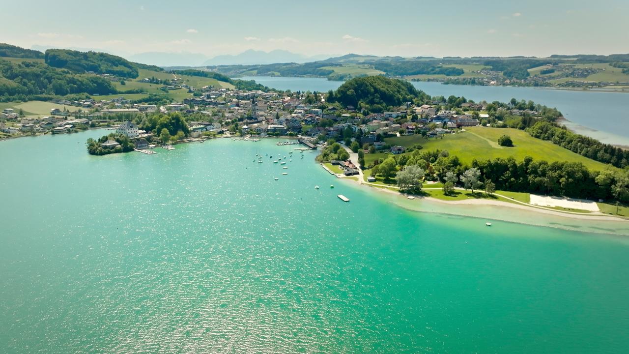 Das Bild zeigt eine weitläufige Landschaft des Mattsees im Salzburgerland, Österreich. Der See ist von klarem, türkisfarbenem Wasser umgeben. Am Ufer sind Schwimmboote und kleine Yachten zu sehen, die sanft im Wasser treiben.   Im Hintergrund erkennt man eine Ansammlung von Häusern, eingebettet zwischen grünen Wiesen und Bäumen. Die Siedlung ist von sanften Hügeln umgeben, die bis zu einem Massiv in der Ferne ansteigen, wo sich auch die Umrisse von Bergen andeuten.   Die Szenerie strahlt eine friedliche, naturnahe Atmosphäre aus, ideal für Erholungssuchende und Wassersportler. Einige Strände sind sichtbar, die zum Entspannen einladen. Das Wetter scheint sonnig und freundlich.