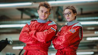 Szene aus "Umlaufbahnen" von Samantha Harvey: Ein junger Astronaut und eine junge Astronautin stehen mit verschränkten Armen nebeneinander. Sie blicken in die Kamera, die Frau trägt eine Brille. Sie sind mit roten Overalls bekleidet. Auf dem rechten Arm des Mannes ist eine russische Flagge zu sehen. Die großbritannische Flagge befindet sich auf dem rechten Arm der Frau. Der Hintergrund ist unscharf, grau und es sind mehrere Leuchtstoffröhren zu erkennen.