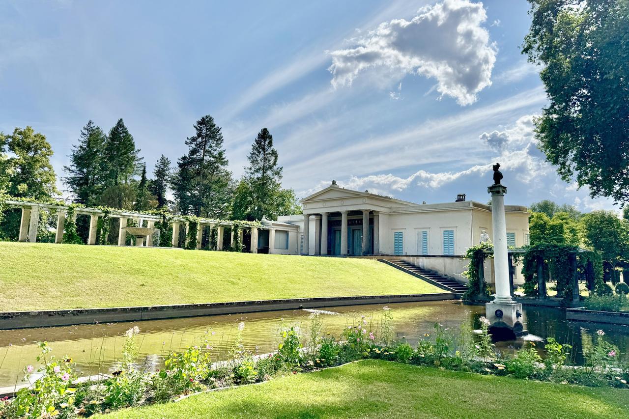 Das Bild zeigt das Schloss Charlottenhof, ein historisches Bauwerk in Potsdam, umgeben von malerischen Gärten. Im Vordergrund erstreckt sich ein kleiner Teich, der die grüne Landschaft widerspiegelt. An den Rändern des Teiches wachsen verschiedene Blumen, die Farben und Texturen zur Szenerie hinzufügen.  Im Hintergrund ist das Schloss sichtbar, das eine klare, architektonisch strukturierte Fassade hat, geschmückt mit Säulen und großen Fenstern mit blauen Fensterläden. Über der Anlage ist der Himmel größtenteils blau mit einigen weißen, wolkigen Elementen. Ein schmaler Weg führt vom Teich zu den Haupttreppen des Schlosses.  Links im Bild steht eine Pergola, die mit Pflanzen bewachsen ist und einen schattigen Bereich bietet. Die gesamte Szenerie strahlt eine ruhige, harmonische Atmosphäre aus, die den Charakter des historischen Ortes unterstreicht.