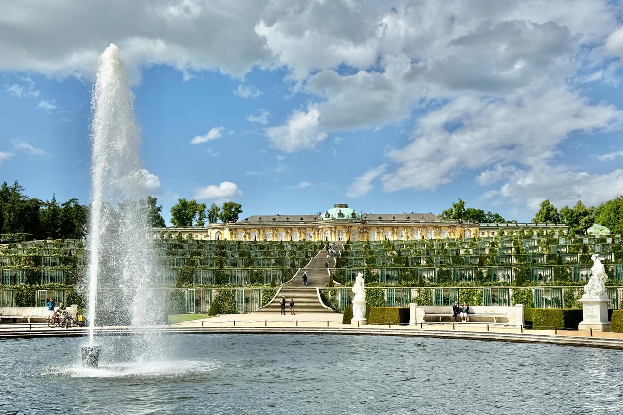 Das Bild zeigt das Schloss Sanssouci in Potsdam, das von weitläufigen barocken Gärten umgeben ist. Im Vordergrund befindet sich ein großer Brunnen, aus dem Wasser in die Höhe sprudelt, umgeben von einem Teich. In der Mitte des Bildes erstreckt sich eine breite Treppe, die zu den terrassierten Gärten führt, die mit Hecken und niedrigem Grün gestaltet sind. Auf den Stufen sind einige Besucher zu sehen, die das Gelände erkunden. Hinter der Treppe steht das Schloss, dessen Fassade aus hellen Steinen besteht und von großen Fenstern und einem auffälligen, kuppelförmigen Dach mit grünem Belag gekrönt wird. Der Himmel ist teils bewölkt, mit hellen Wolken, die über das Gelände ziehen. Insgesamt vermittelt das Bild einen Eindruck von historischer Architektur in einem gepflegten Gartenumfeld.