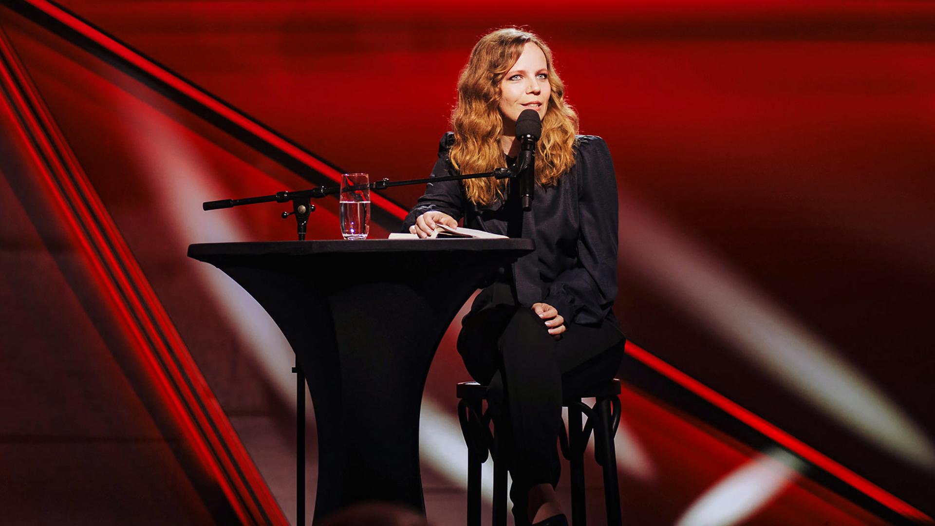 Sarah Bosetti sitzt auf einem Barhocker. Links von ihr ist ein Stehtisch. Die Bühne ist düster und rot beleuchtet.