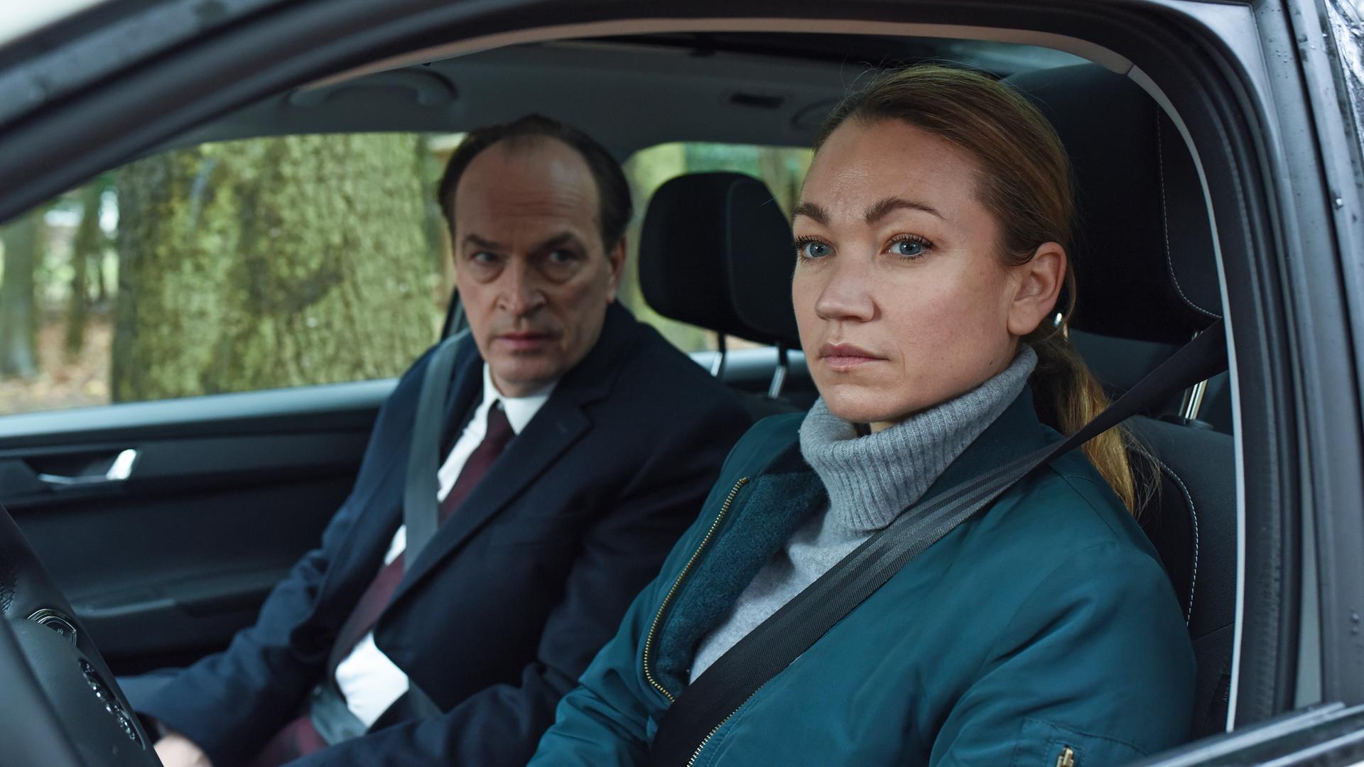 Eine Frau sitzt am Steuer eines Wagens, auf dem Beifahrersitz ein Mann, er sagt etwas zu ihr. Beide wirken nachdenklich, angespannt. 