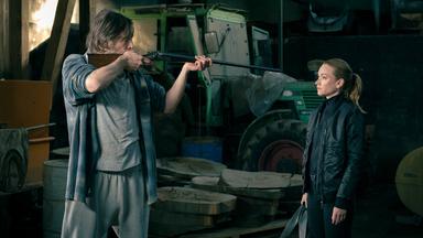 Axel Thal (Alexander Wüst) richtet in einer dunklen Scheune ein Gewehr auf Sarah Kohr (Lisa Maria Potthoff); im Hintergrund sind landwirtschaftliche Geräte und Holzscheite sichtbar.