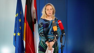 Eine Frau steht vor zwei Mikrofonen und redet. Im Hintergrund stehen Fahnen.