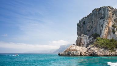 Fotografie vom Strand Cala Luna auf Sardinien: Türkisblaues Mittelmeer trifft auf steile Felsklippen, im Hintegrund ein kleines Boot auf dem Wassser.